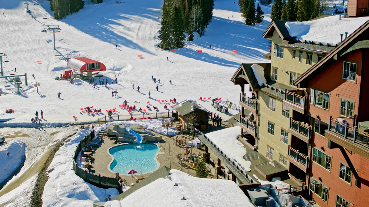 A vibrant exterior showing the lodge's pool area surrounded by snow, active skiers on the slopes, and après-ski gatherings beside the building. The juxtaposition of heated pool and snowy landscape highlights both relaxation and adventure.
