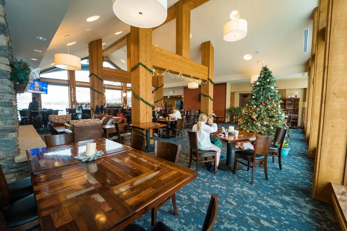 A warm, inviting lodge interior featuring large wooden pillars, a festive Christmas tree, cozy seating areas, and a welcoming bar. The space is lively yet relaxing, ideal for gathering after a day of mountain adventure.