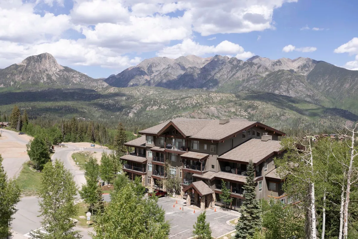 This exterior image showcases a three-story lodge set against a dramatic mountain backdrop, surrounded by lush green landscapes typical of summer in the San Juans. The scene hints at hiking and nature walks starting right from the property, with inviting outdoor areas perfect for relaxing after adventurous days.