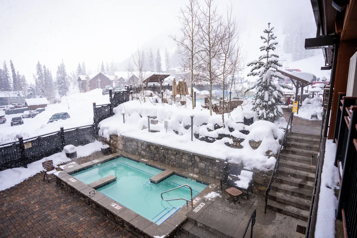 A tranquil scene featuring an outdoor hot tub surrounded by snow, with trees and steps leading down to the relaxation area. Guests soak in the tub, enjoying mountain views and the quiet magic of a snowy evening.