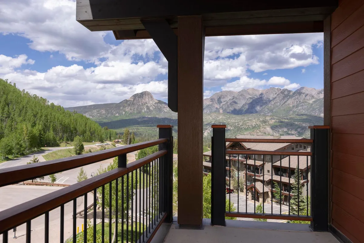 The image showcases the exterior balcony of a luxury condo overlooking a breathtaking mountain range, with lush greenery all around. The spacious balcony features a sturdy railing, perfect for lounging and soaking in the peaceful surroundings. The scene evokes tranquility, with the promise of sunrise views, stargazing at night, and the possibility of spotting wildlife in the nearby forest.