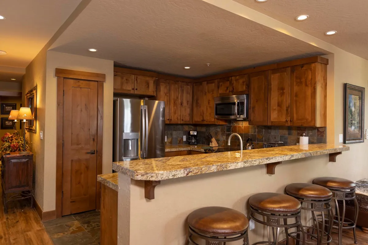 A modern, well-equipped kitchen with granite countertops, stainless steel appliances, wood cabinetry, and bar stools. The kitchen flows seamlessly into the adjacent living space, inviting both cooking and socializing.