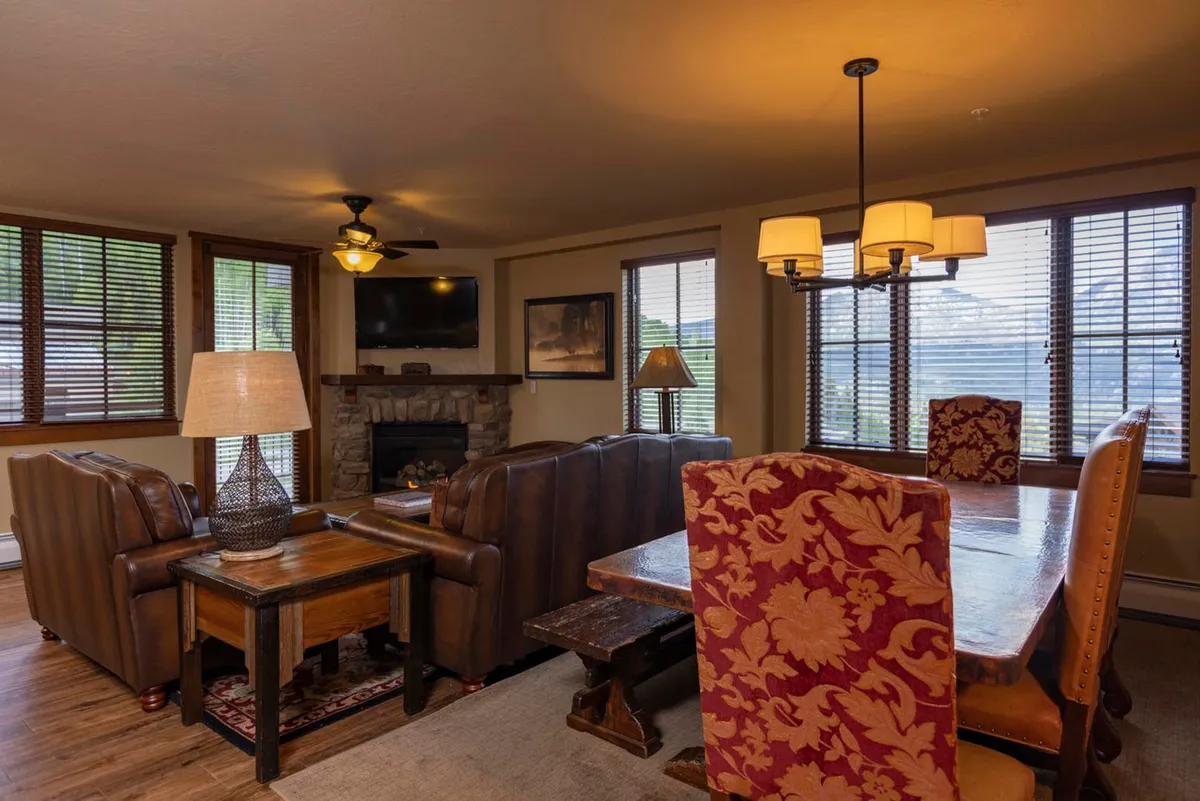 A spacious open-plan living and dining area featuring plush leather sofas arranged around a stone fireplace, a large dining table set nearby, and expansive windows that flood the space with natural light. The windows offer glimpses of mountain views beyond, and the overall ambiance is warm and inviting, perfect for group gatherings.