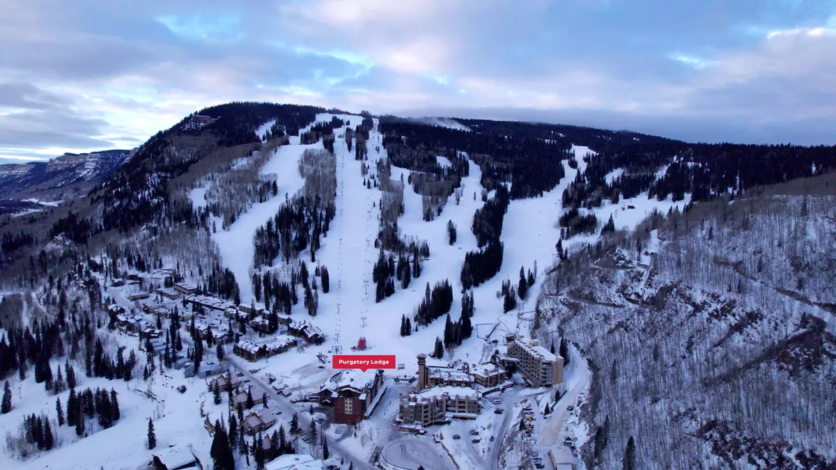 A sweeping winter exterior of Purgatory Lodge nestled at the base of snow-covered ski slopes, with majestic mountains and snow-laden trees framing the lodge. The scene captures skiers and snowboarders gliding down the slopes, with the warmth of the lodge visible in the background.