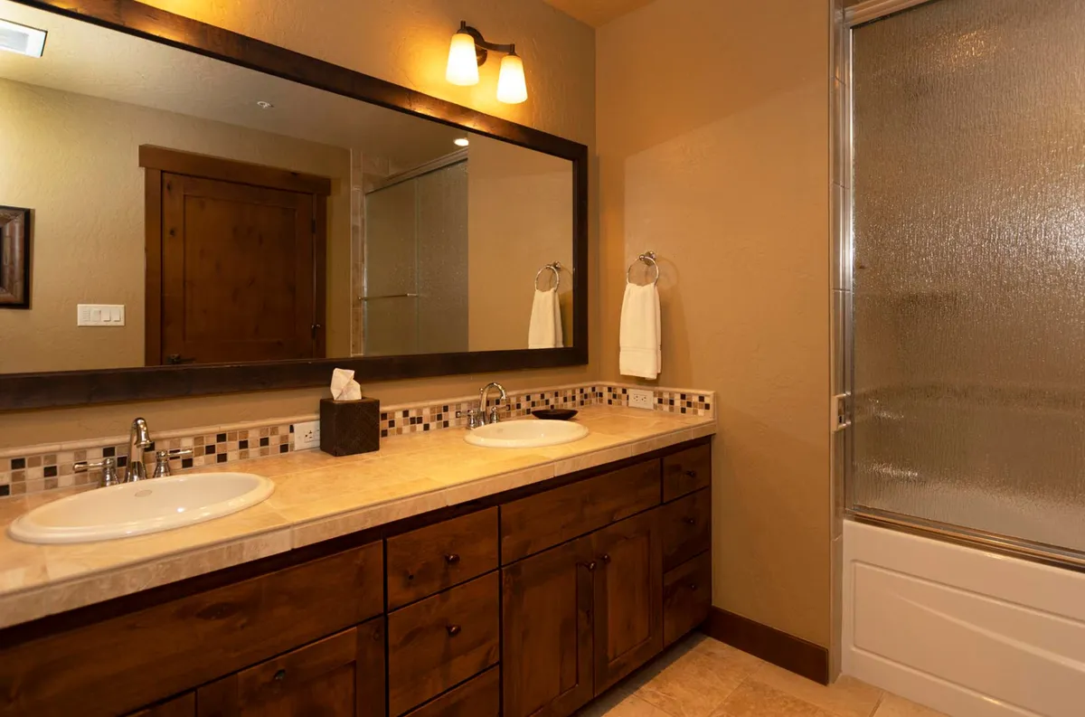 A serene bathroom with a double sink vanity, expansive mirror, and a glimpse of the shower area. The tiled backsplash and clean lines create a spa-like ambiance perfect for rejuvenation and self-care.