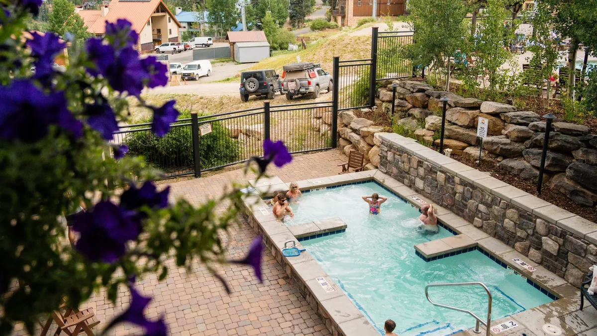 An outdoor swimming pool surrounded by vibrant flowers, with families enjoying time in the pool and hot tub. The scene is lively and captures both socializing and relaxation. The pool area appears inviting and well-maintained, suggesting a premium experience.