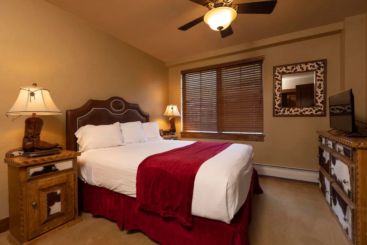 A cozy bedroom featuring a comfortable bed adorned with a vibrant red throw blanket, flanked by a wooden nightstand and a decorative mirror. Soft natural light streams through the window with blinds, creating a warm and inviting atmosphere ideal for unwinding after a day of adventure.