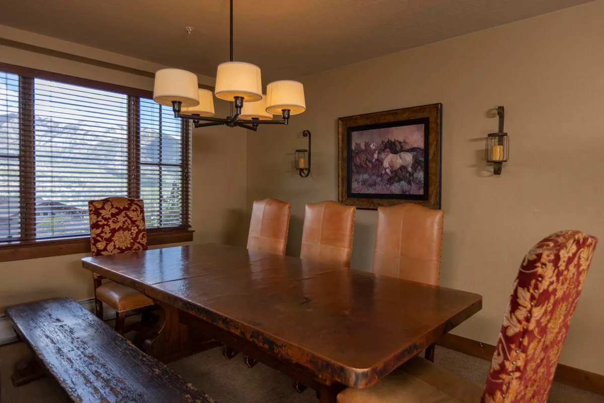 A cozy dining space with a large wooden table surrounded by upholstered chairs, a statement chandelier, and tastefully selected artwork. Natural light streams through the windows, offering serene views and a welcoming atmosphere.