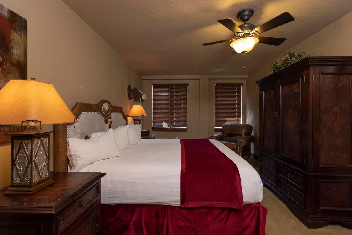 A serene king bedroom with plush white bedding, a classic wooden armoire, decorative lamp on a nightstand, and a comfortable armchair by the window. The setting invites relaxation and peaceful moments in a restful atmosphere.