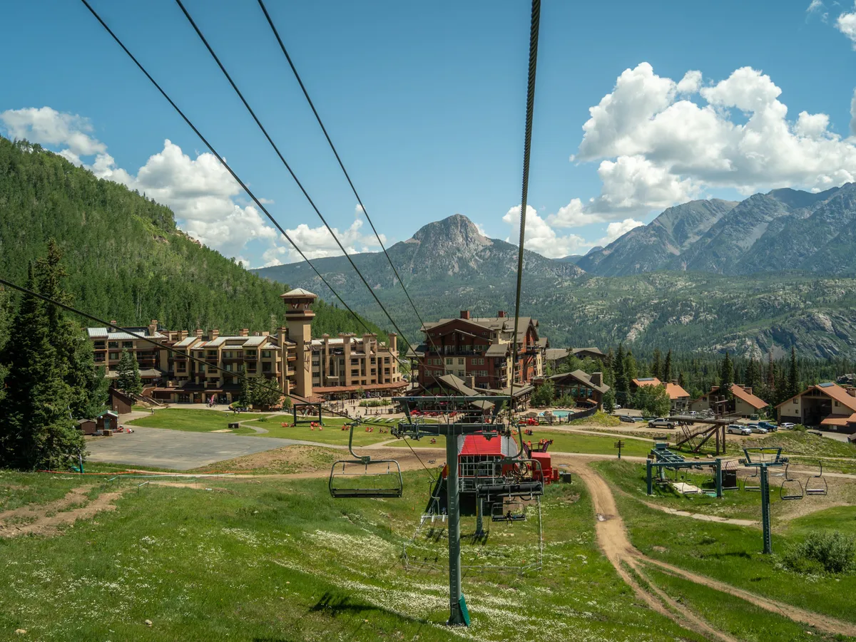Featuring ski resort buildings, lush green grass, and a gondola lift set against a mountain backdrop, this exterior shot emphasizes the range of activities possible in summer, from hiking and mountain biking to simply soaking in serene views. The gondola hints at chairlift-accessed biking and hiking mentioned in the text.