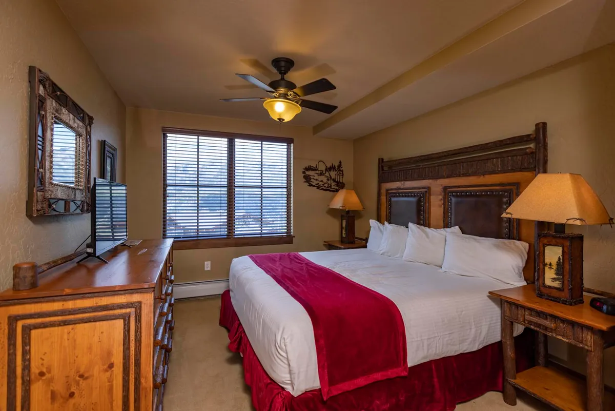 A bright, tranquil bedroom featuring a king-sized bed with crisp white linens, a wooden dresser, bedside lamps, and a large window with blinds letting in natural light. The room exudes comfort and a subtle mountain-inspired style, ideal for relaxation after an active day.
