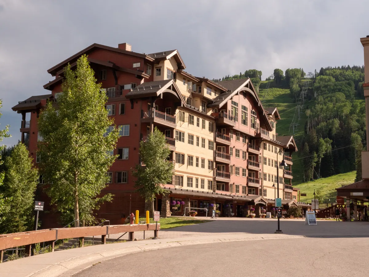An animated exterior scene with a multi-story building nestled against a lush green hillside, featuring outdoor seating and shop signage. The area exudes vibrancy, suggesting access to both mountain adventures and village amenities.